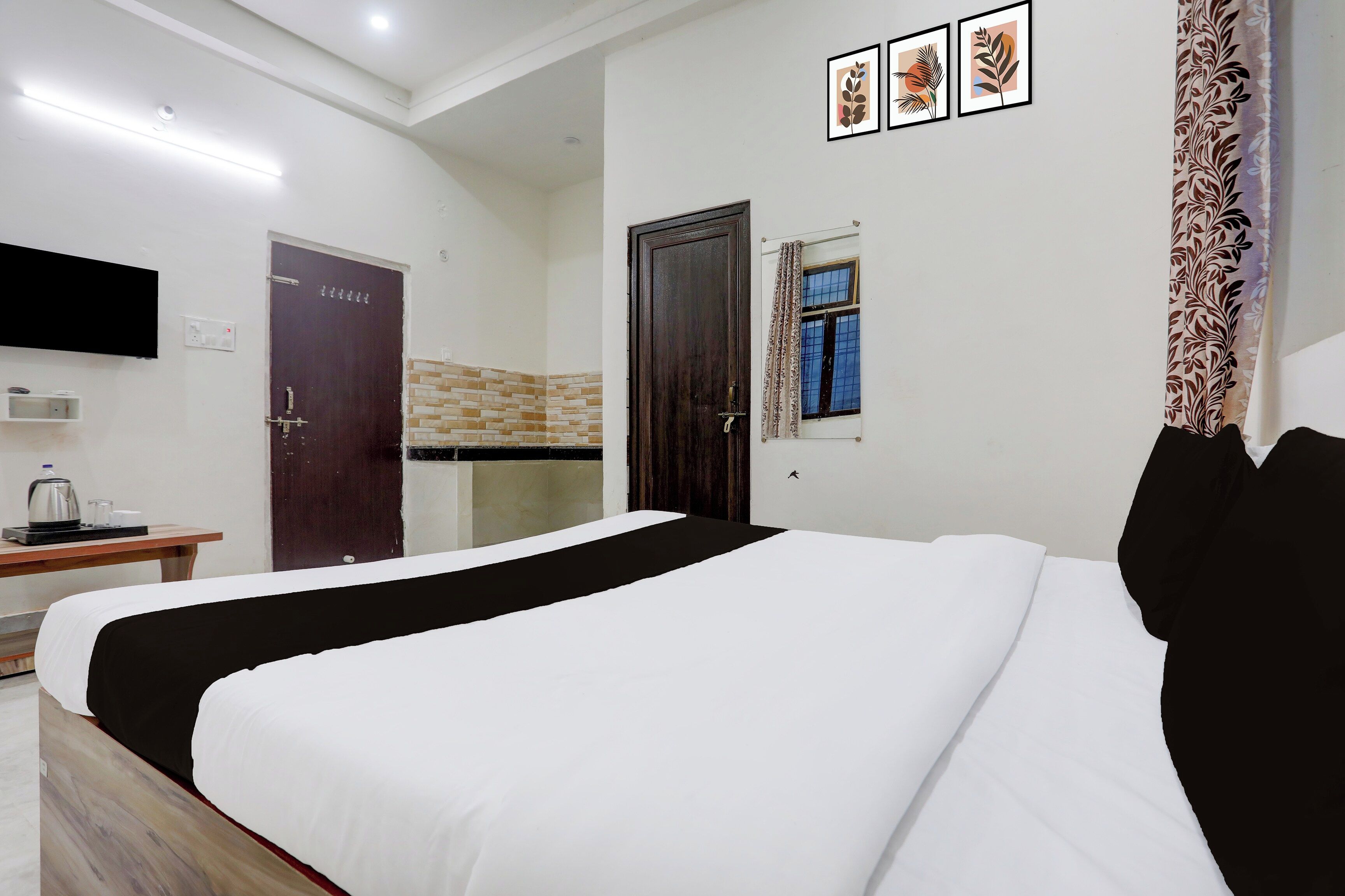 A hotel room with a bed, a desk with a kettle, a TV, and a window with curtains.
