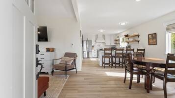 A modern kitchen with white cabinets, a dining area with wooden chairs, and a TV mounted on the wall.