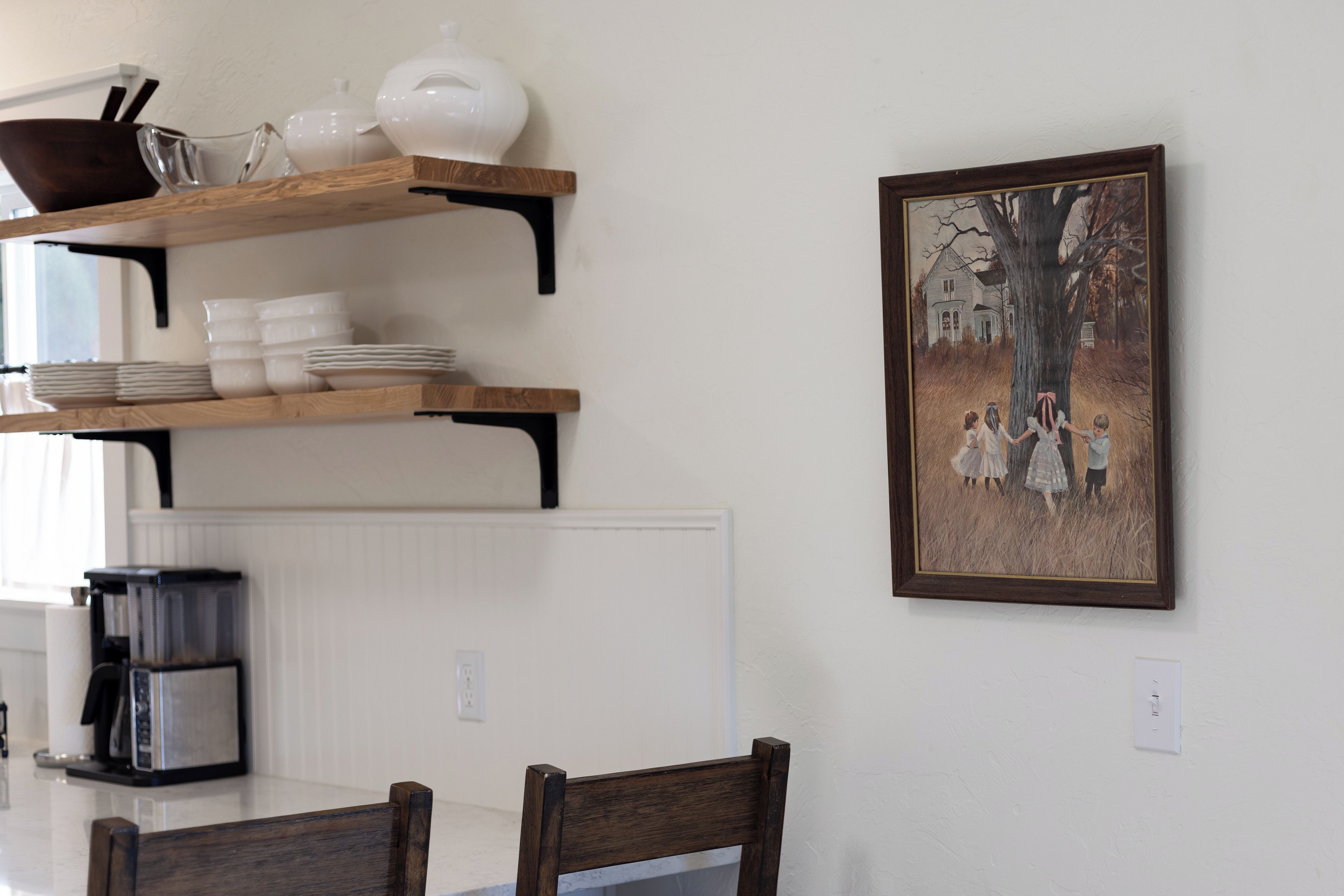 A dining area with a white table, wooden chairs, a coffee machine, and a shelf with dishes and a white pot.