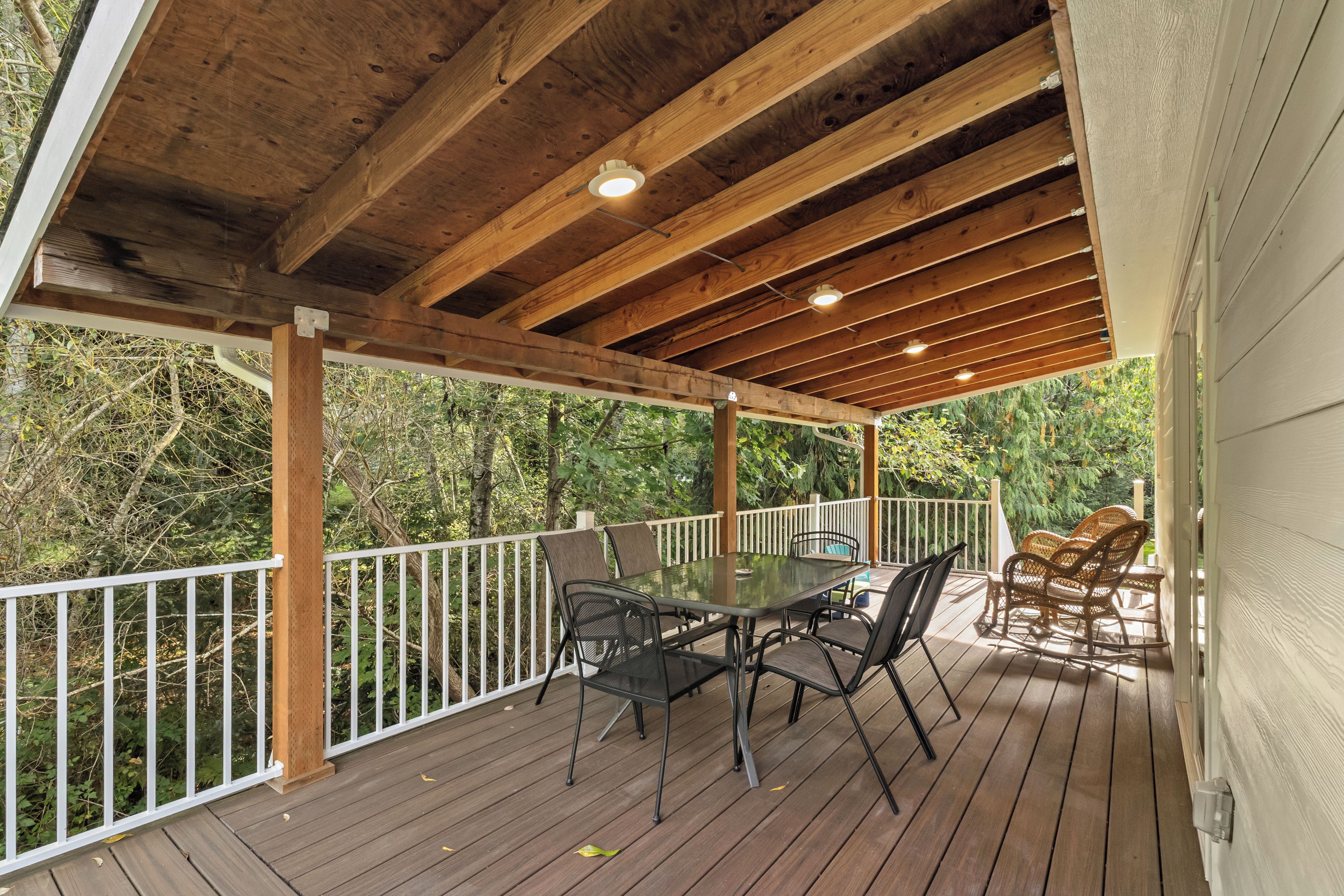 A covered outdoor deck with a table and chairs, surrounded by a white railing, overlooking a wooded area.