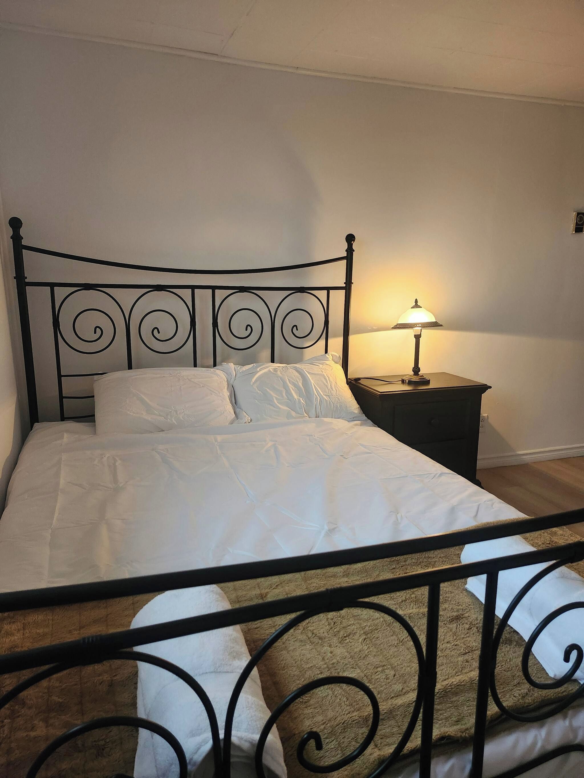 Una cama con estructura metálica, ropa de cama blanca y una mesita de noche con lámpara.