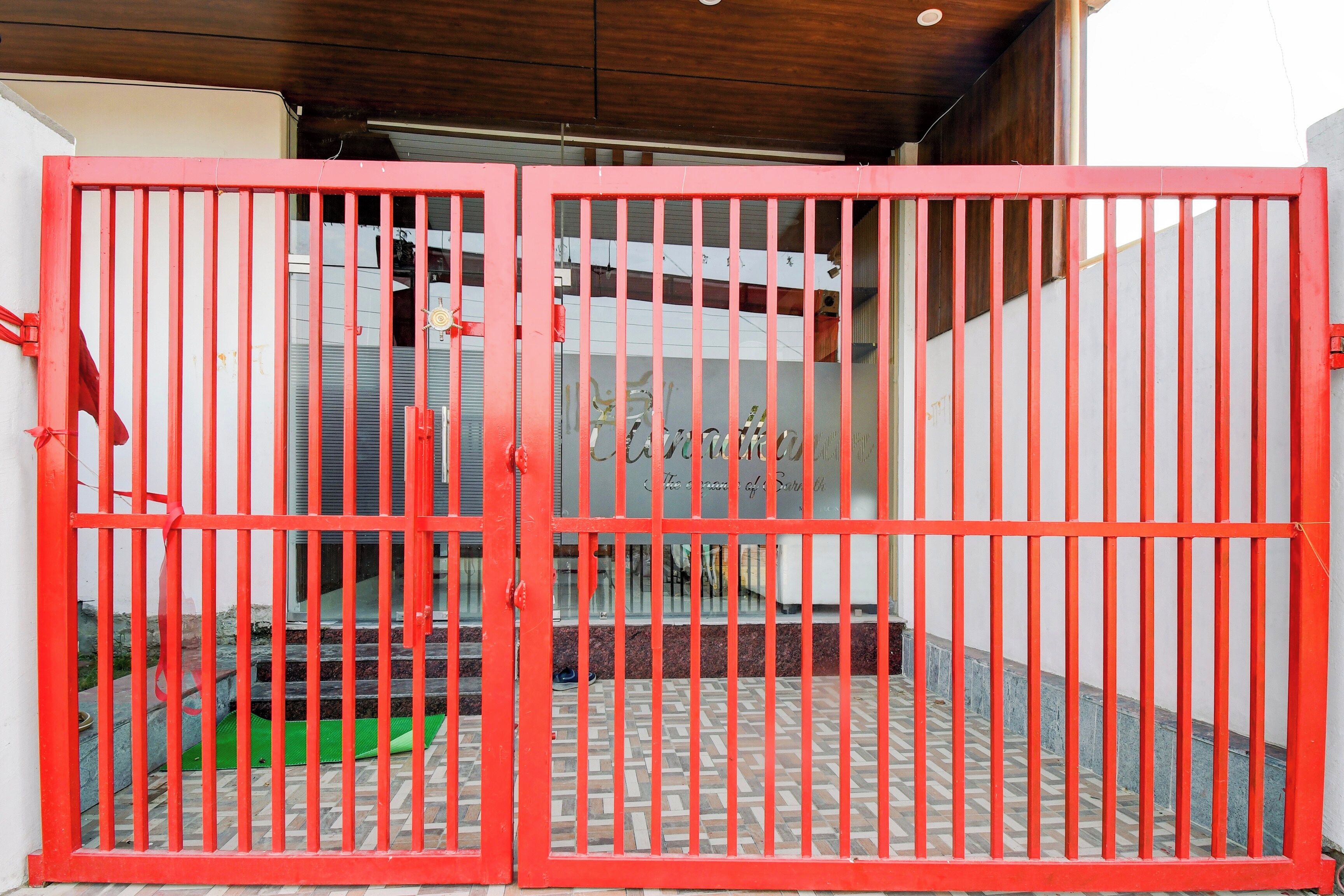 Une porte métallique rouge à barreaux verticaux, un sol carrelé et un mur blanc avec une fenêtre.