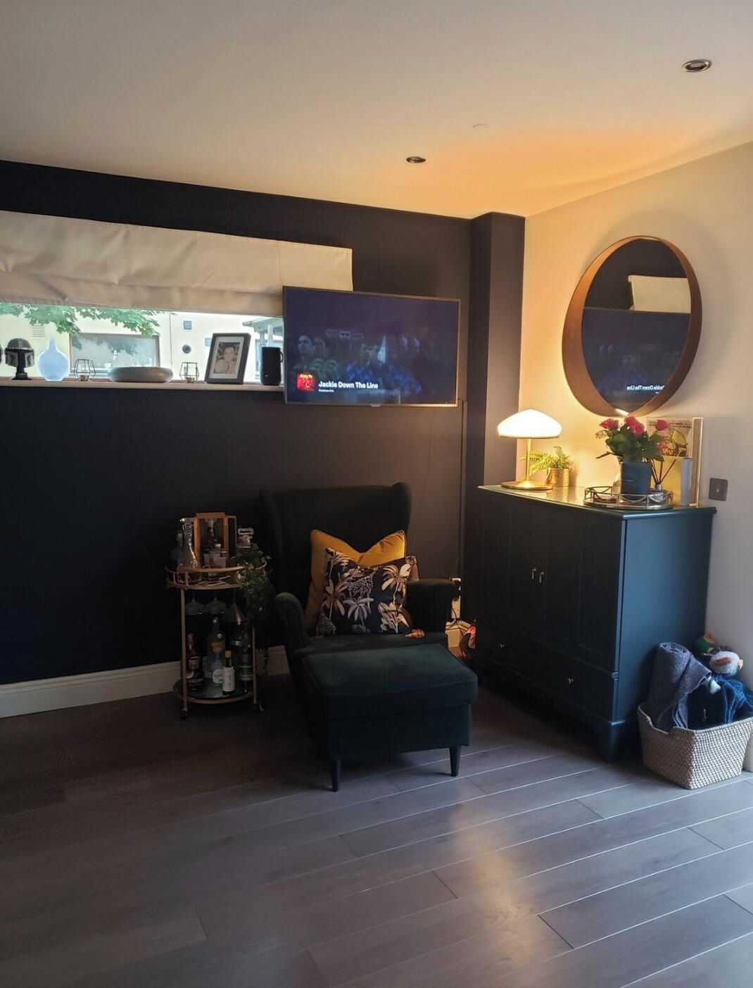 A living room with a dark-colored sofa, a small table with a lamp, a TV on the wall, and a cabinet with a mirror.