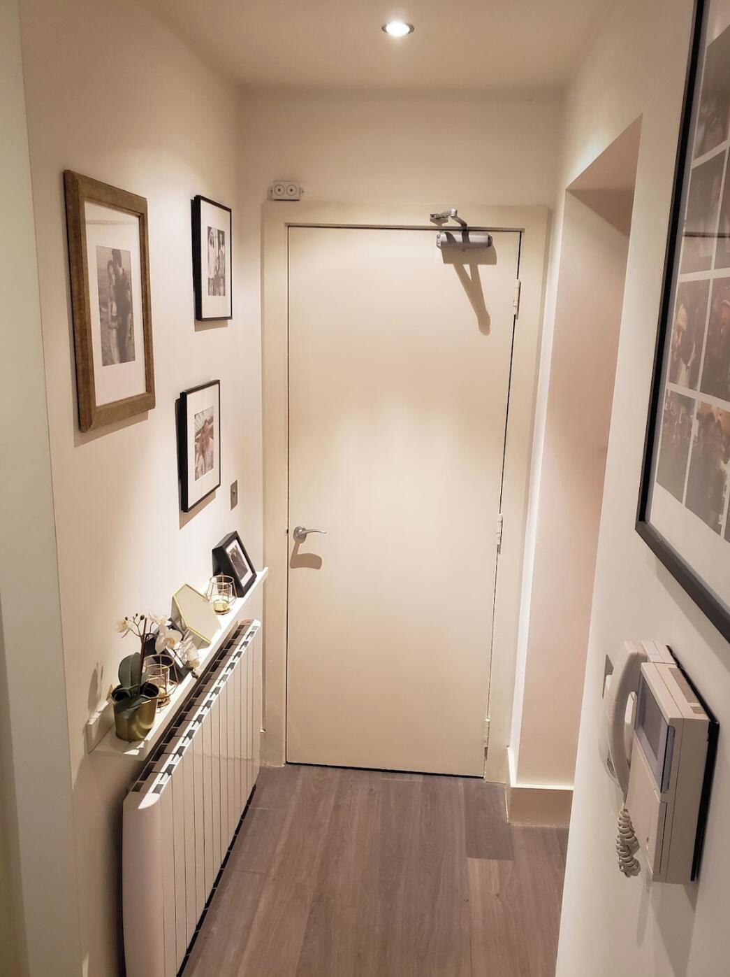 Un pasillo con una puerta blanca, cuadros en la pared, un radiador y un termostato.