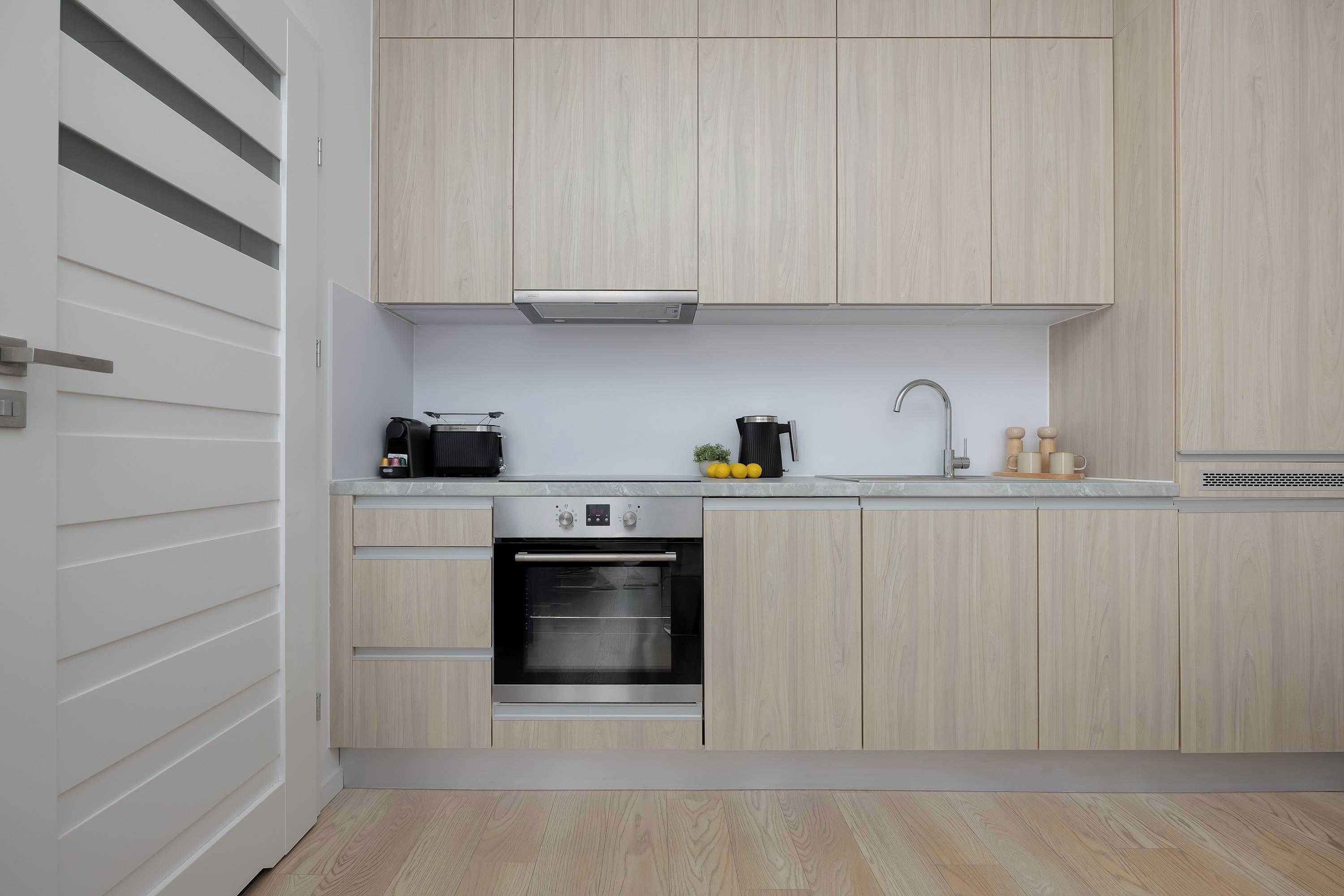 A modern kitchen with light wood cabinetry, a built-in oven, a coffee maker, and a sink.