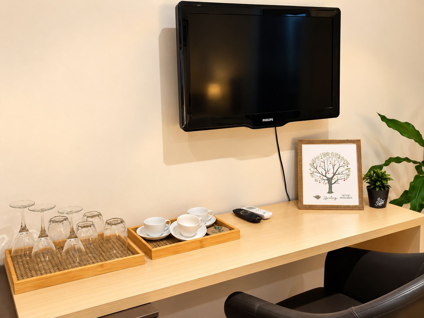 A room with a flat-screen TV mounted on the wall, a wooden desk with a tray of wine glasses, cups, and a framed picture.