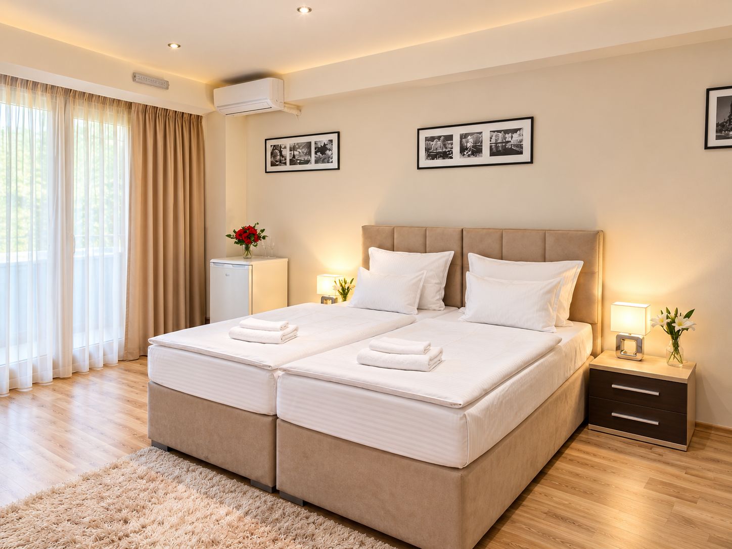 A double bed with a headboard, bedside tables with lamps, a small refrigerator, and a large window with curtains.