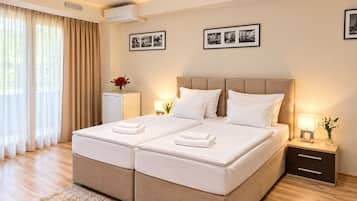A double bed with a headboard, bedside tables with lamps, a small refrigerator, and a large window with curtains.