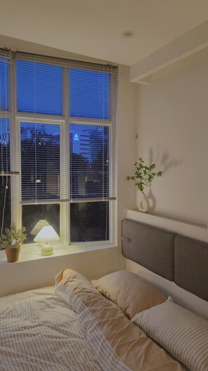 A bedroom with a bed, a window with blinds, a bedside lamp, a potted plant, and a wall-mounted shelf. - D1 Heart · Cityview & Rooftop · Kanso @ Cozinema (Ben Thanh, District 1)