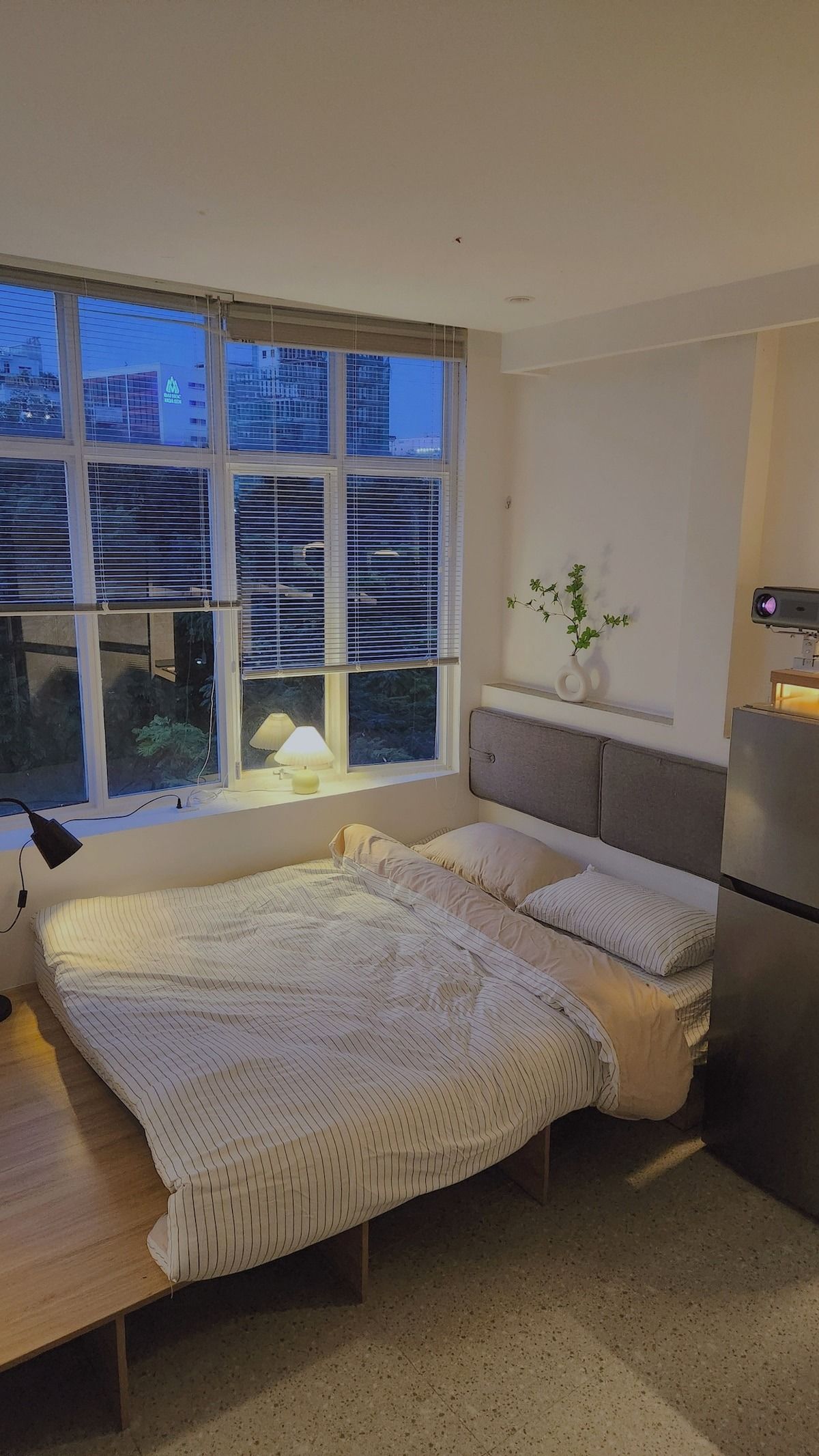 A bedroom with a bed, a desk, a lamp, a plant, and a window with blinds.