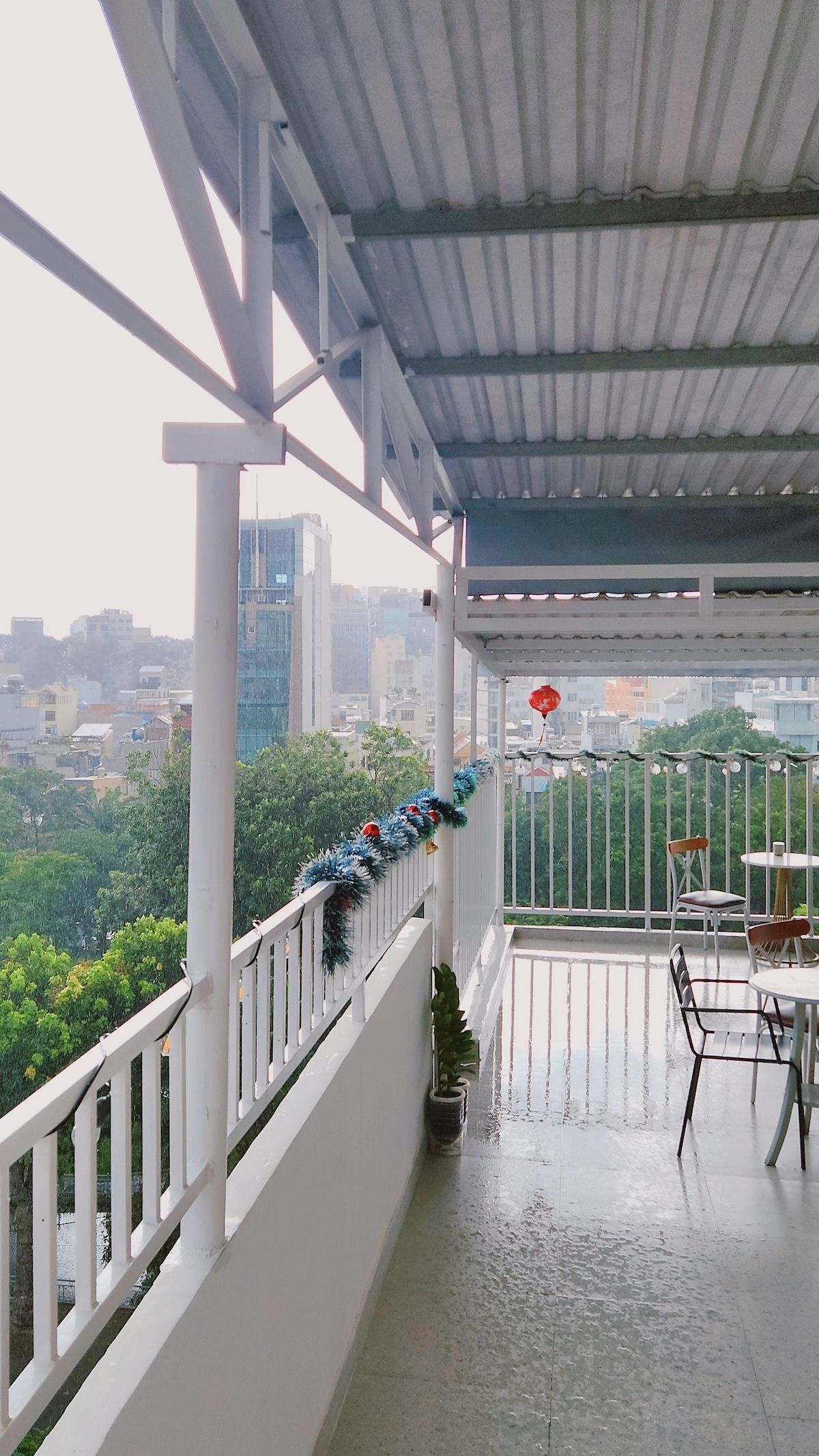 A covered balcony with a view of a cityscape, featuring a railing, a potted plant, and a table with chairs.