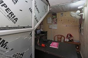 Een compact kantoor met een bureau, stoel, computer en diverse decoraties, waaronder een ventilator en religieuze posters.