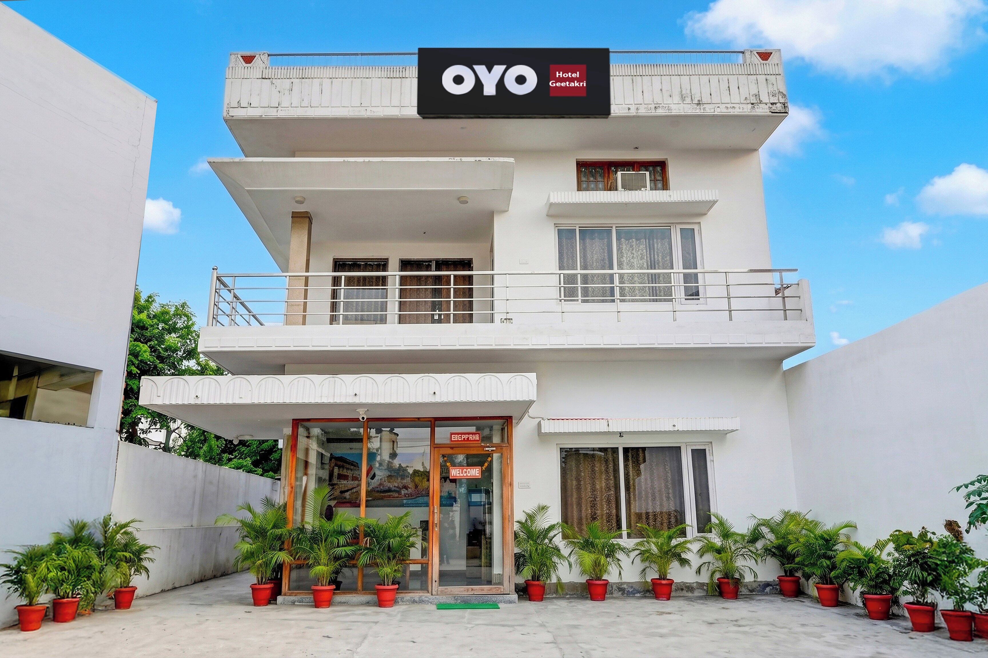 Ein zweistöckiges Hotel namens 'OYO Hotel Geetakri' mit einem Balkon im oberen Geschoss, einem Glastür-Eingang und Blumentöpfen im Vordergrund.