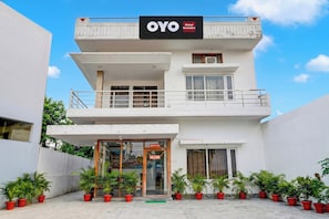 Un hotel de dos pisos llamado 'OYO Hotel Geetakri' con un balcón en el piso superior, entrada de vidrio y plantas en macetas al frente.
