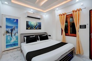 Une chambre d’hôtel avec un lit, un appareil de climatisation, une porte ornée d’un tableau représentant un paysage marin, et une fenêtre avec des rideaux.