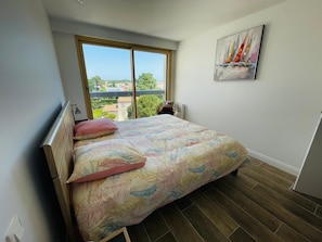 Quarto com uma grande janela, cama com roupa de cama floral, um quadro na parede e vista para o exterior.