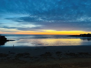 Uma praia com mar calmo, uma pessoa caminhando e um pôr do sol ao fundo.