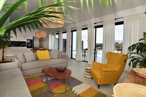 A modern living room with a sofa, armchair, and coffee table, a kitchen area, and large windows with curtains. - New Listing (La Oliva)