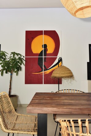 Un coin repas avec une table en bois, des chaises en osier, une plante en pot et une œuvre d’art murale représentant la silhouette d’une personne avec un soleil en arrière-plan.