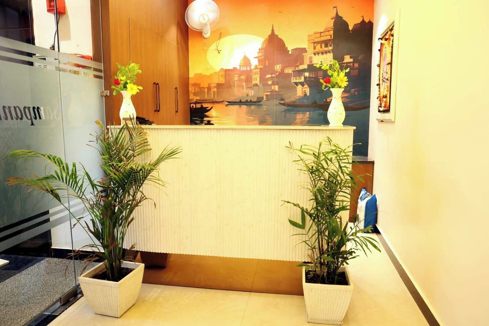 Un mostrador de recepción con un mural de paisaje urbano, dos plantas en macetas y un ventilador encima.
