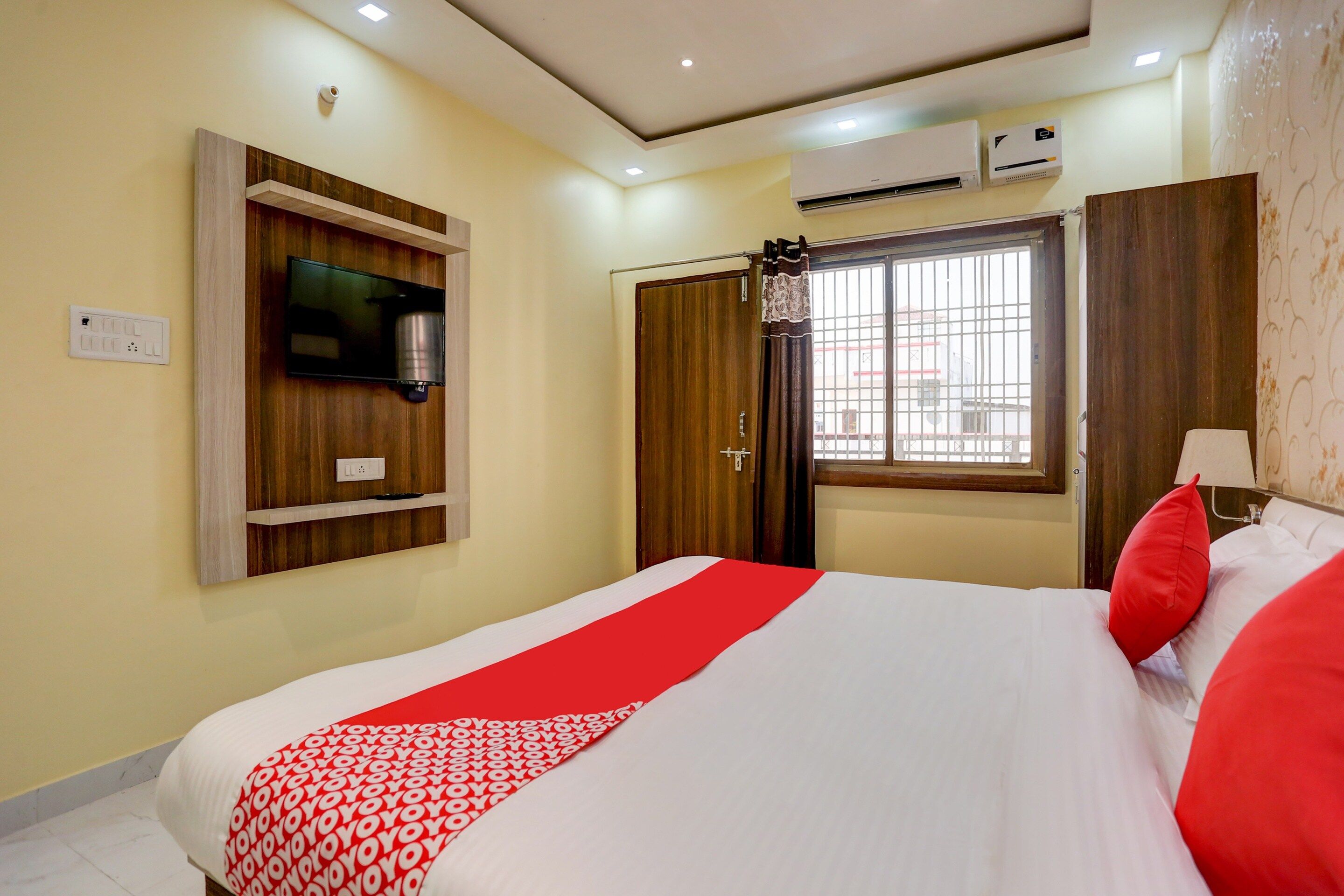 A hotel room with a bed, a television mounted on the wall, an air conditioning unit, and a window with a view of buildings outside.