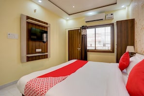 Habitación de hotel con cama, televisor montado en la pared, equipo de aire acondicionado y ventana con vista a edificios.