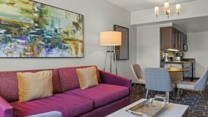 Una habitación de hotel moderna con un sofá morado, una mesa de comedor con sillas, una lámpara de pie y un cuadro abstracto grande en la pared.