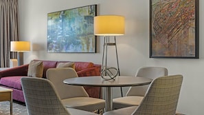 Quarto de hotel com uma mesa redonda, duas cadeiras, um sofá e duas luminárias de chão.
