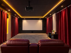 Sala de cine en casa con una pantalla grande, cortinas rojas, asientos de cuero y un proyector.