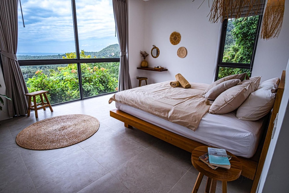 Une chambre avec un lit, un petit tabouret, un tapis rond en fibres naturelles et une vue sur la verdure grâce à de grandes fenêtres.