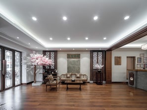 Área de recepção com balcão de madeira, espaço de estar com sofás e cadeiras, plantas decorativas e um tapete de parede tradicional.