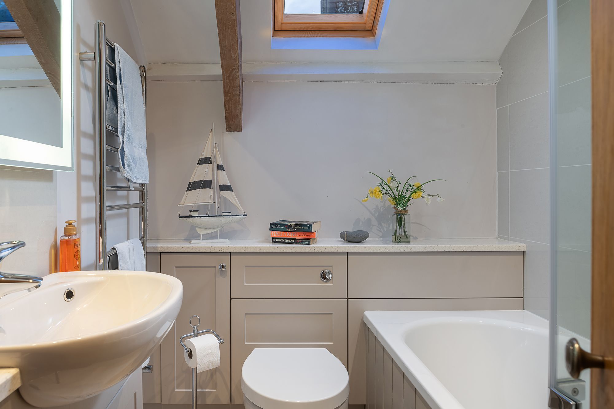 Un baño con un lavamanos grande, inodoro y bañera. Hay una ventana pequeña sobre el lavamanos, un perchero y un estante con libros y un florero con flores.