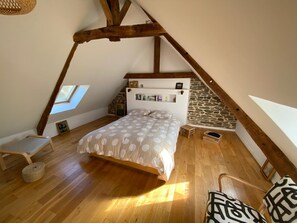 Un dormitorio con techo inclinado, vigas de madera, una cama con edredón de lunares, un taburete de madera, una silla y una pared de piedra.
