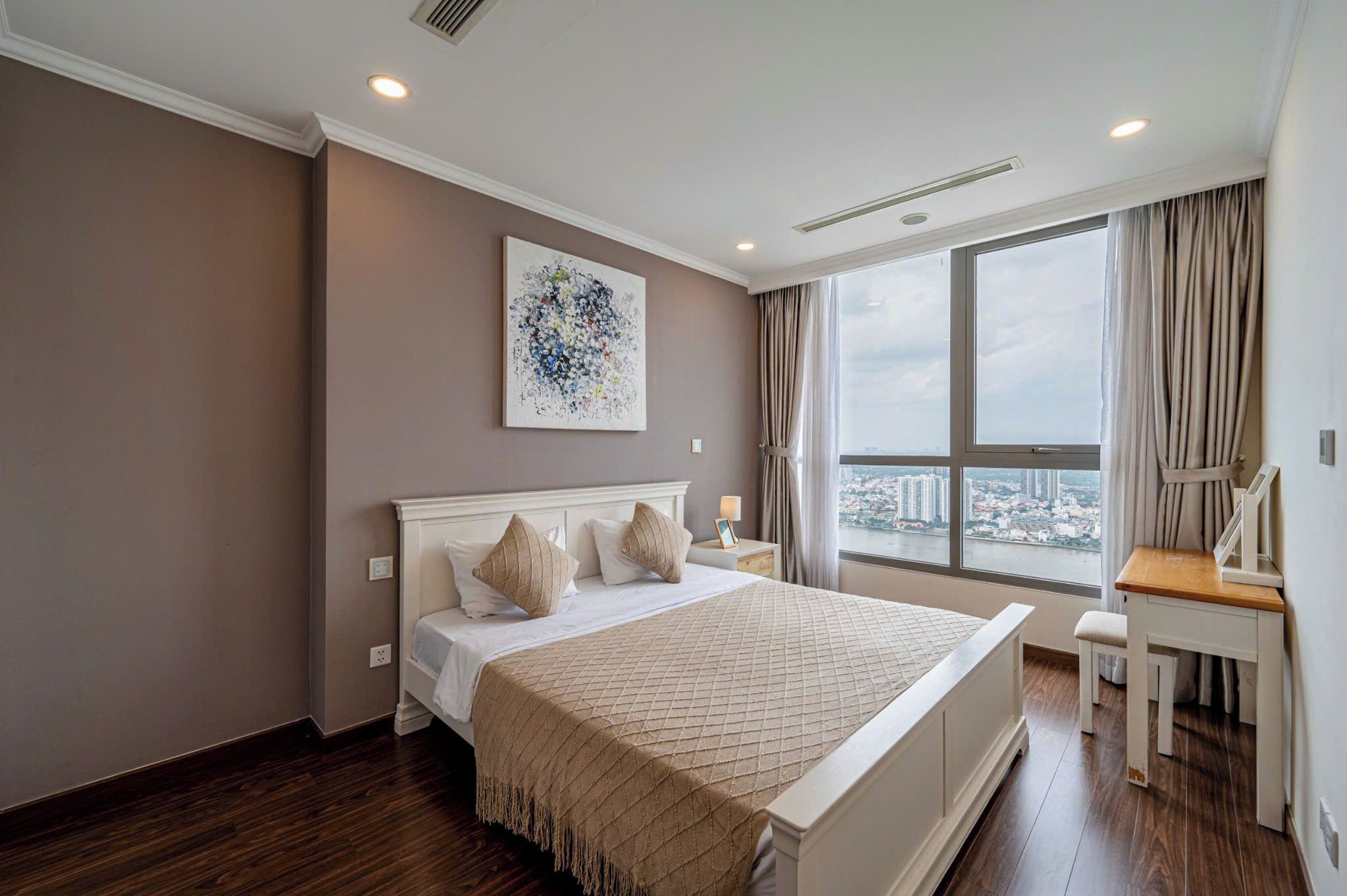A hotel room with a large bed, a desk, a chair, a window with a city view, and a ceiling light.