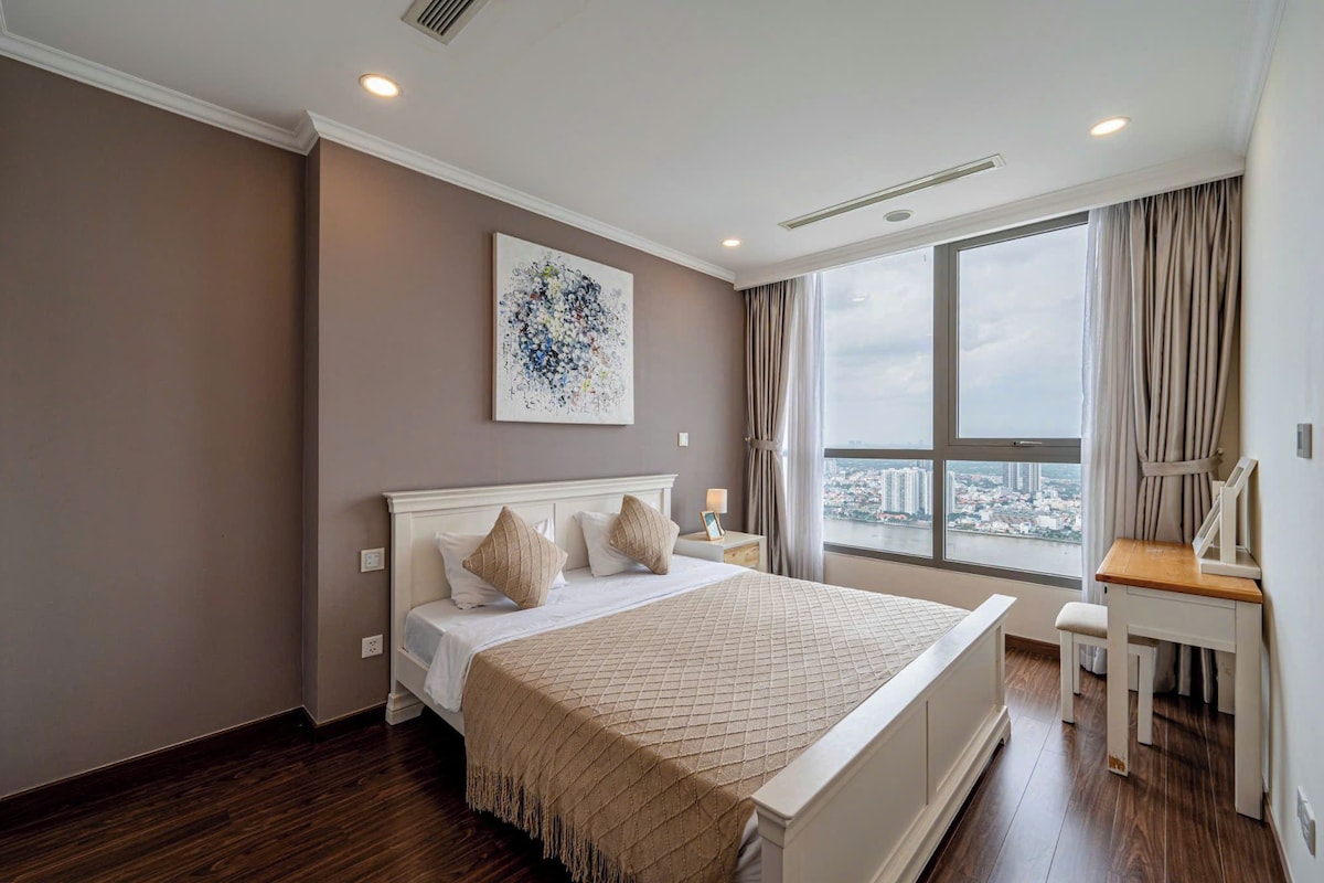 Quarto de hotel com cama grande, escrivaninha, cadeira, janela com vista para a cidade e luminária no teto.