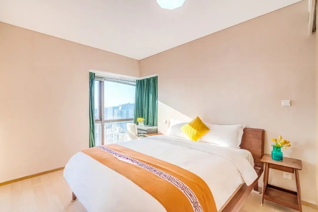 Ein Hotelzimmer mit einem großen Bett, einem Nachttisch, einem Fenster mit Vorhängen und Blick auf die Stadt.