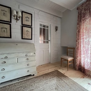 Een kamer met een witte ladekast, een stoel, een deur met een raam en ingelijste foto's aan de muur.