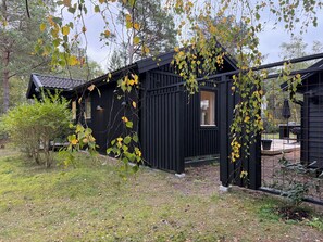Eine schwarze Holzhütte mit einer kleinen Veranda und einem Fenster, umgeben von Bäumen und Gras.