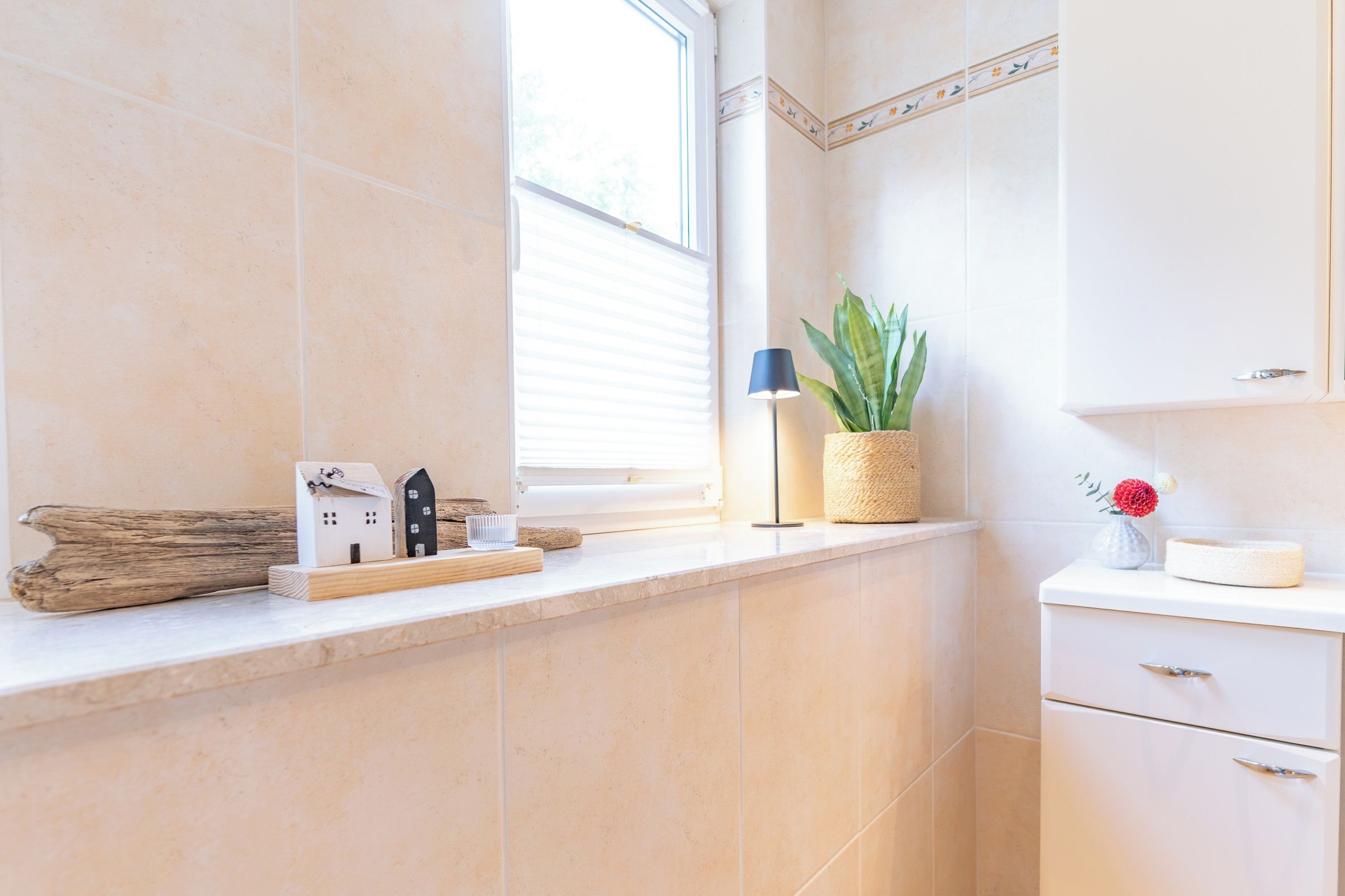 Un baño con ventana, una planta, una lámpara y un pequeño modelo de casa sobre el mostrador.