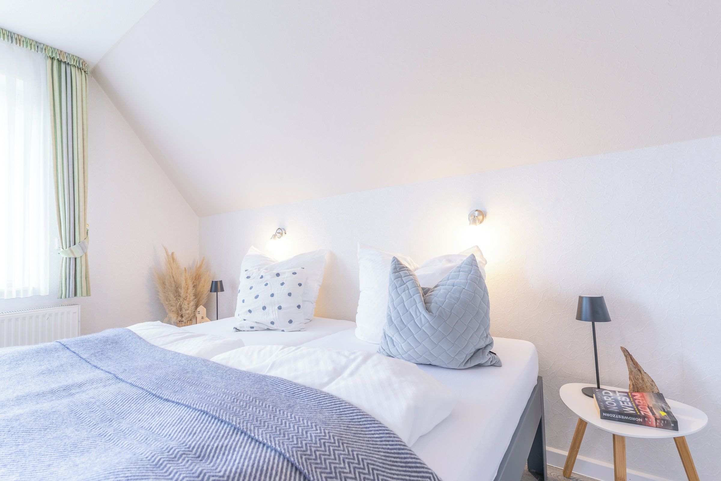 A bedroom with a bed, pillows, a bedside table with a lamp, and a window with curtains.