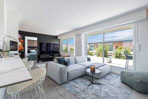 Una sala moderna con un sofá gris, un comedor con sillas blancas, un televisor de pantalla plana y ventanas amplias con vistas a la ciudad.