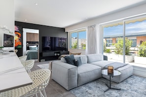 Un salon moderne avec un canapé gris, un coin repas avec des chaises blanches, un téléviseur à écran plat et de grandes fenêtres offrant une vue sur la ville.