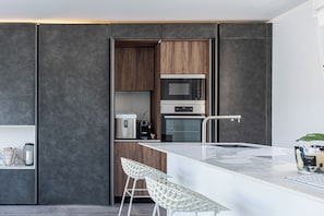 Una cocina moderna con microondas, horno y cafetera integrados. Hay dos taburetes blancos y una encimera de mármol blanco.
