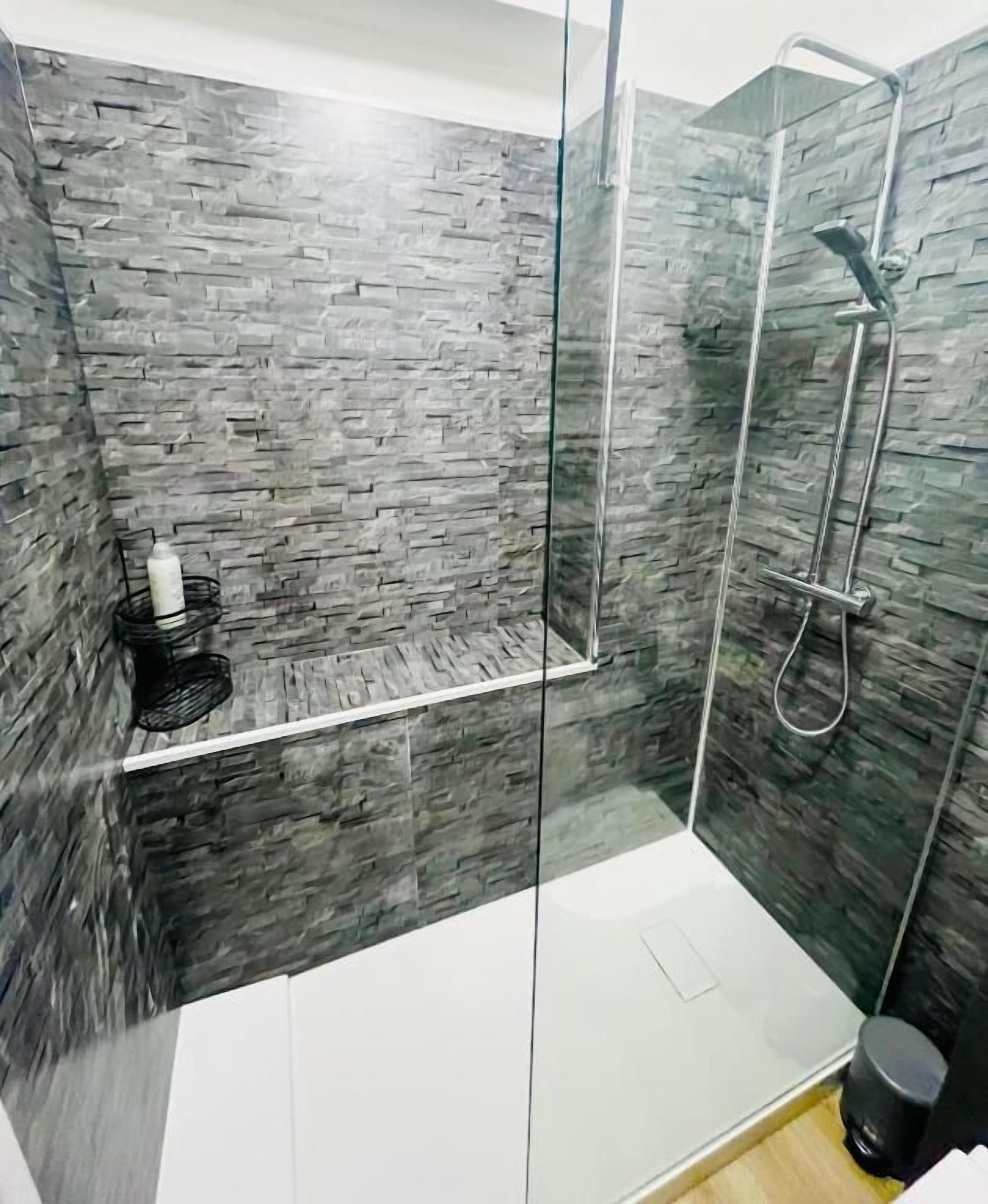 Eine gläserne Dusche mit einer Wand aus Steinoptik, einer Handbrause und einer kleinen Ablage mit einer Flasche.