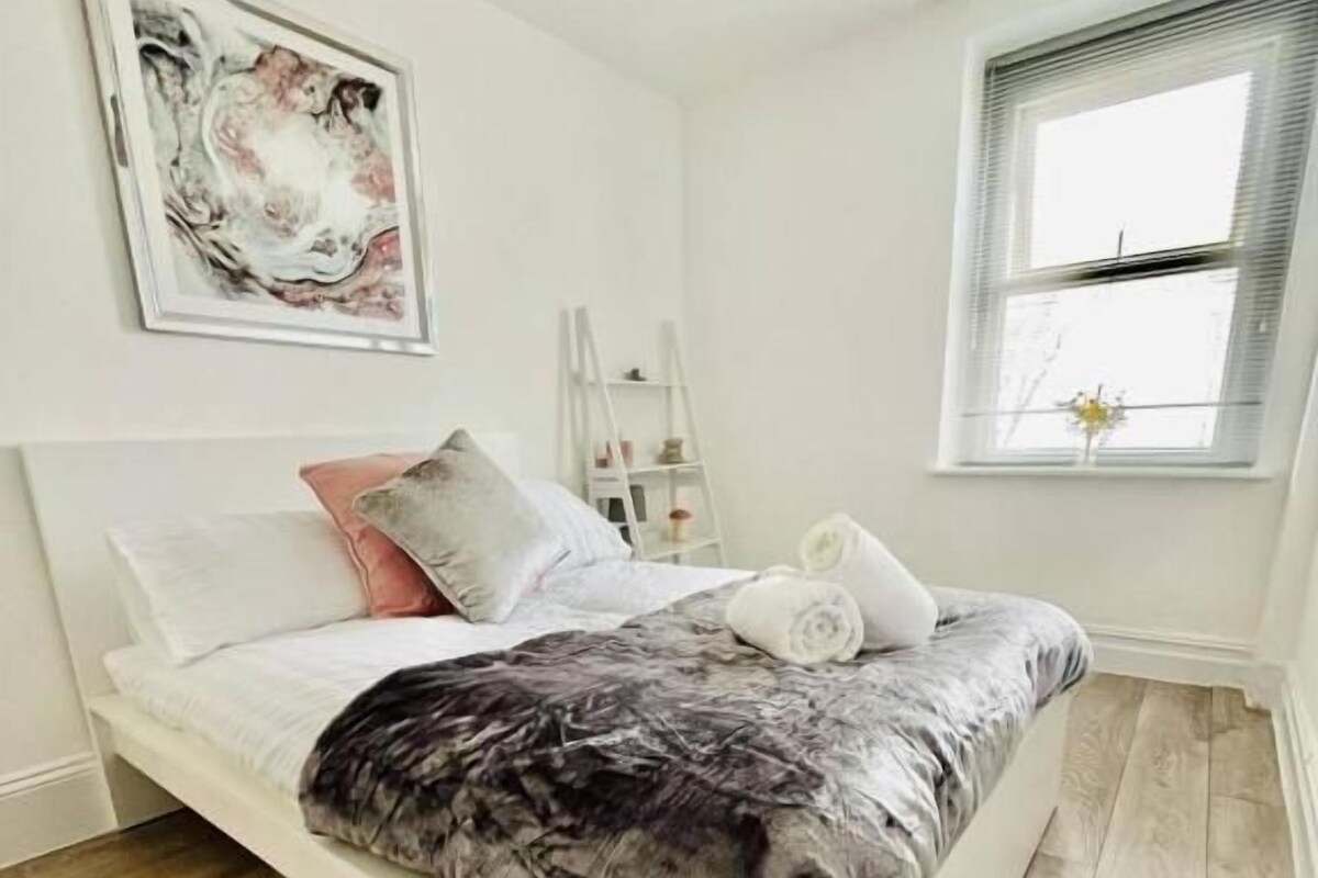 Ein Schlafzimmer mit einem weißen Bett, Holzboden, einem Fenster mit Jalousien und einem Gemälde an der Wand.