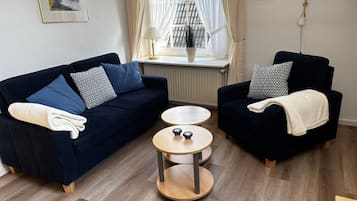 A living room with a dark blue sofa, a matching armchair, a round wooden coffee table, and a window with sheer curtains.