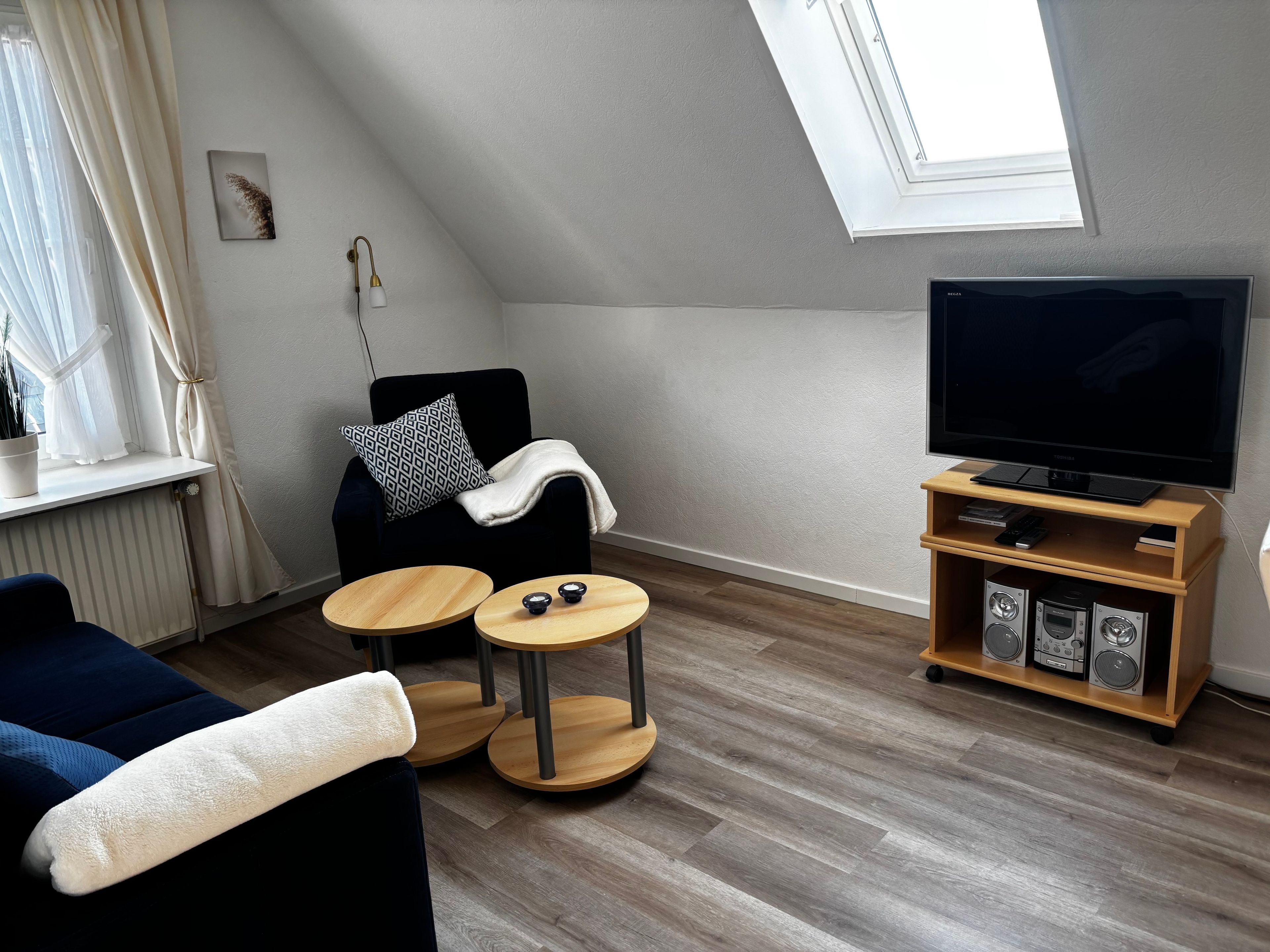 A modern living room with a black sofa, a white throw blanket, a small wooden table with two bowls, a black TV on a wooden stand, and a radiator.
