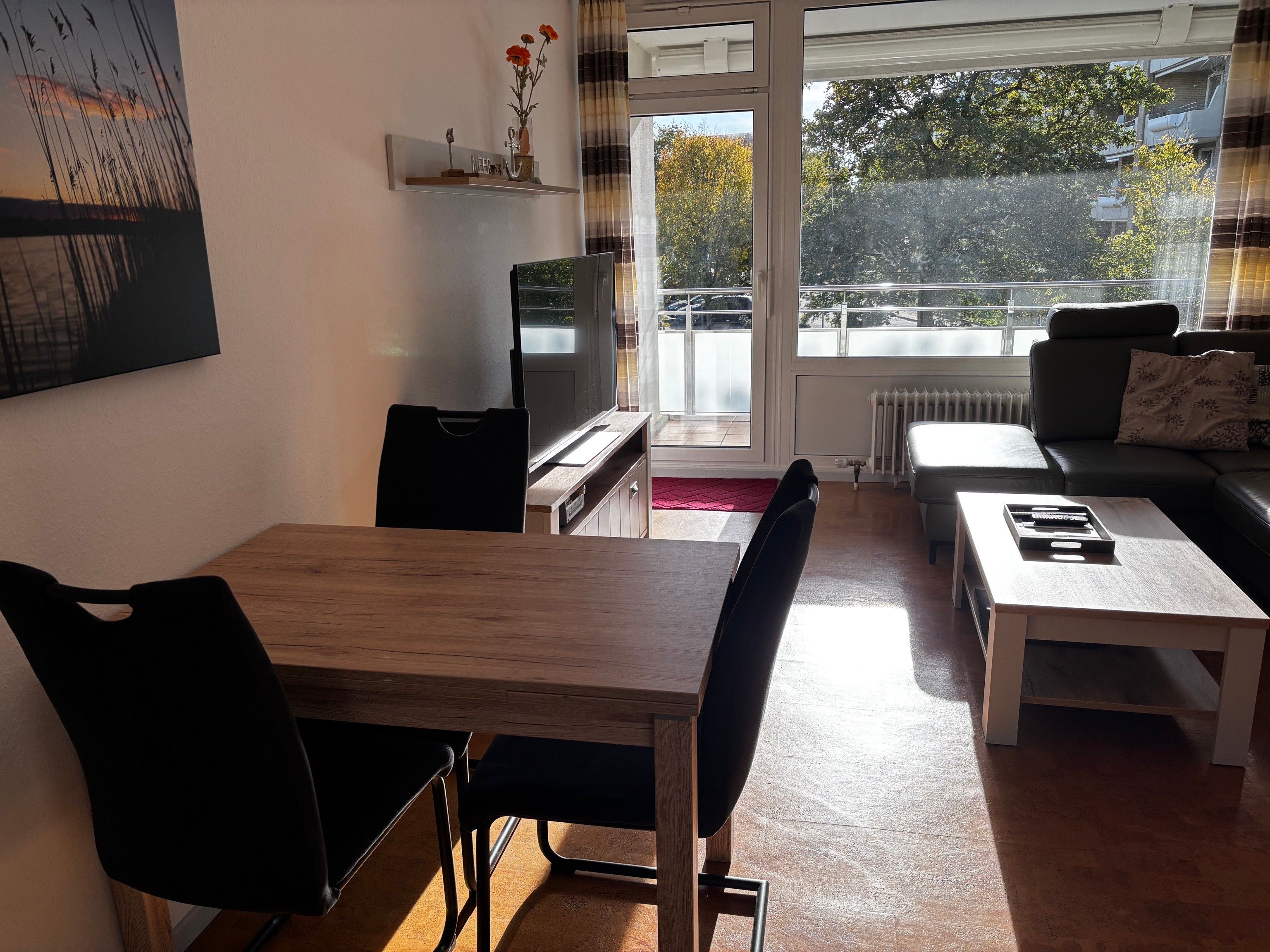 En stue med spisebord, stole, sofa, fjernsyn og et vindue med udsigt til træer og bygninger.