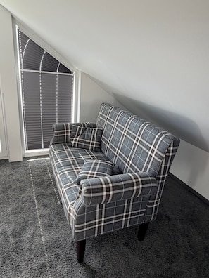 A corner sofa with a plaid pattern in a room with a sloped ceiling and a window with blinds.