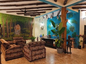 Un vestíbulo con mostrador de recepción, sofás de estampado de leopardo, un amplio mural de un bosque y decoración con plantas tropicales.